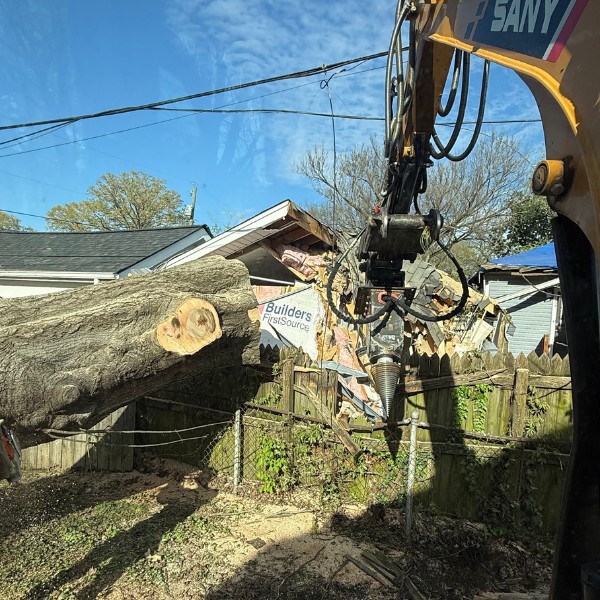 Grapple saw crane lifting a fallen tree trunk near a storm-damaged fence and building